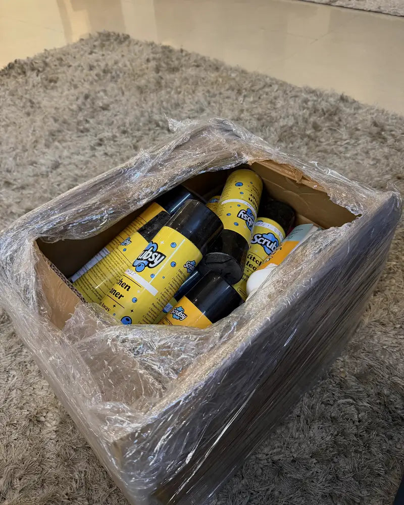 An opened carton of Scrubsy Kitchen Cleaner cans on a rug at home